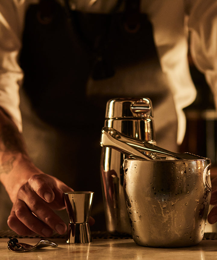Bartender in apron prepares cocktail with stainless steel shaker, jigger, and ice bucket on wooden bar under warm lighting.