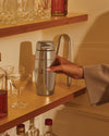 Sleek silver stainless steel cocktail shaker by Alessi, held by a hand on a wooden bar shelf.