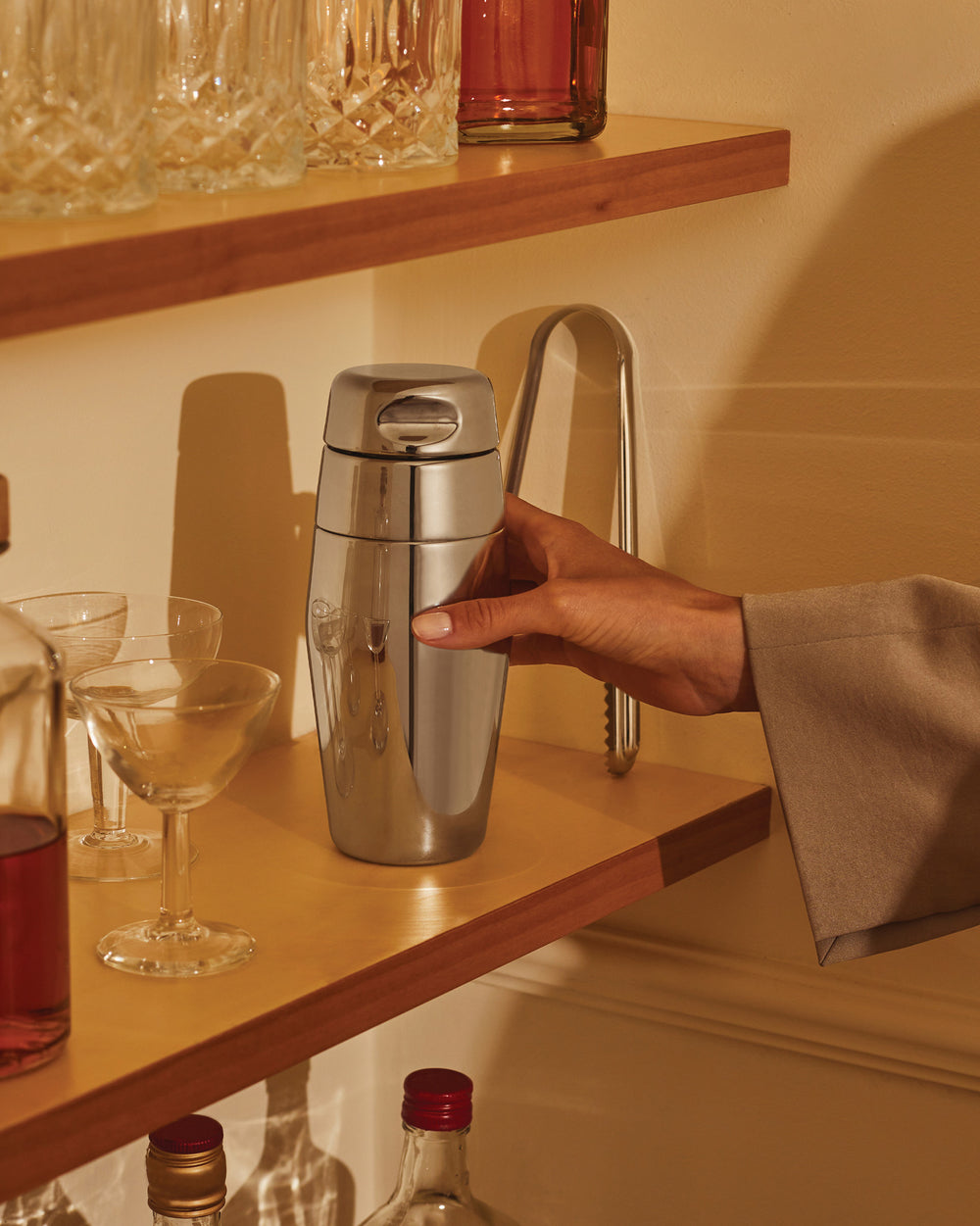 Sleek silver stainless steel cocktail shaker by Alessi, held by a hand on a wooden bar shelf.