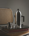 Silver-toned espresso coffee maker by Alessi, featuring a dome top and black handle, on a tray with a small jug and book.