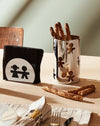 Black napkin holder by Alessi, with a white circular design featuring two stylized cutout figures, on a table.
