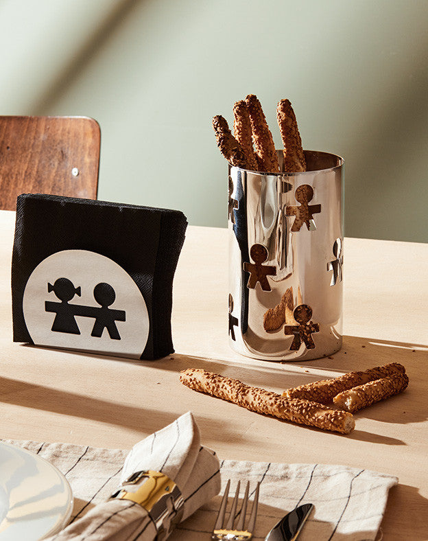 Black napkin holder by Alessi, with a white circular design featuring two stylized cutout figures, on a table.