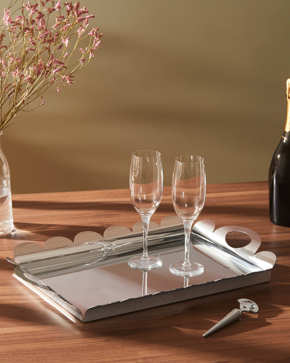 Two clear glass champagne flutes by Alessi, resting on a metallic scalloped tray next to a champagne bottle.