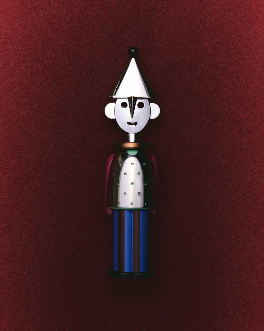 Silver and blue figure corkscrew by Alessi, with a conical hat, perforated torso, and striped base, centered on a dark red background.