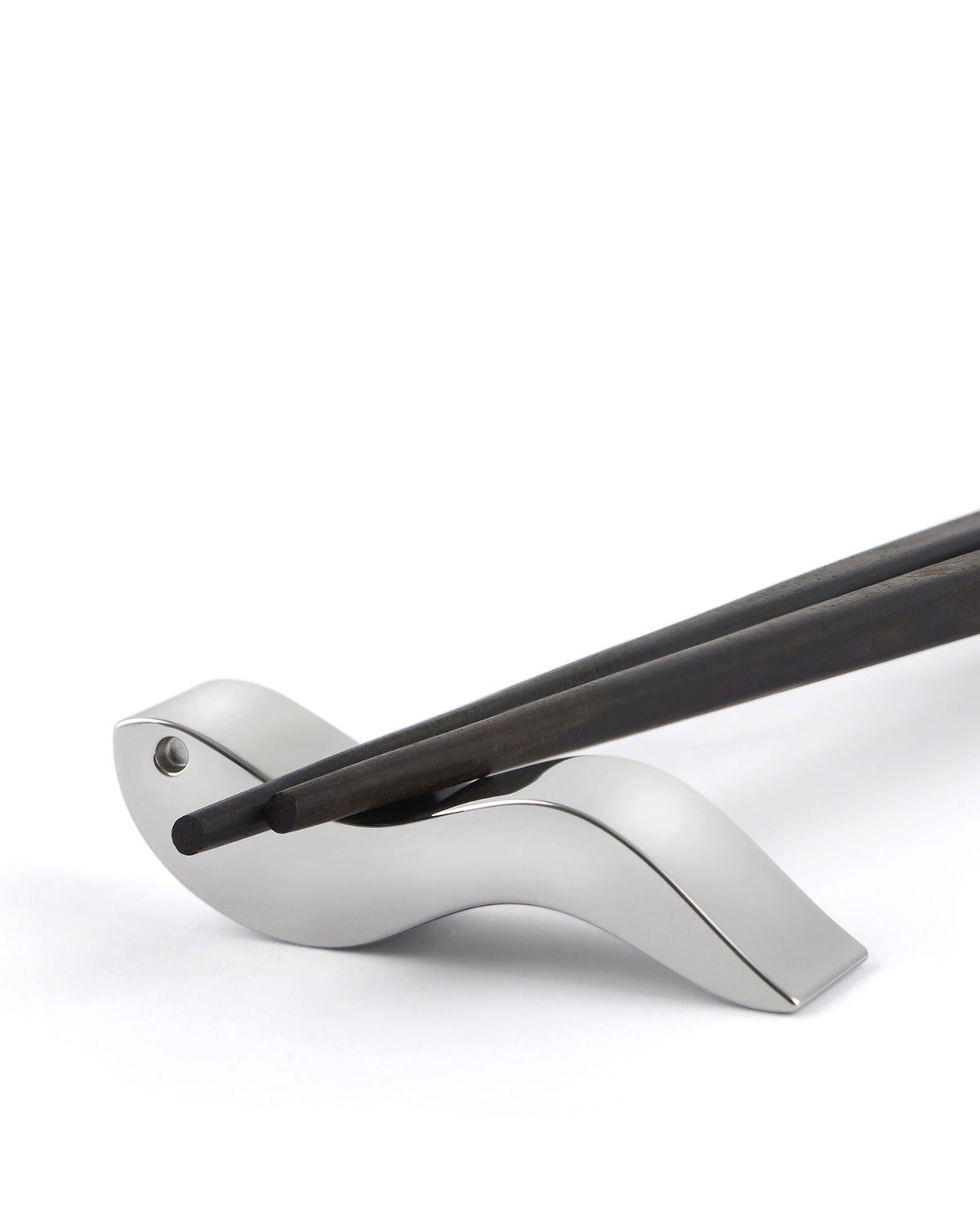 Silver metallic chopstick rest by Alessi holding two dark wooden chopsticks on a white background.