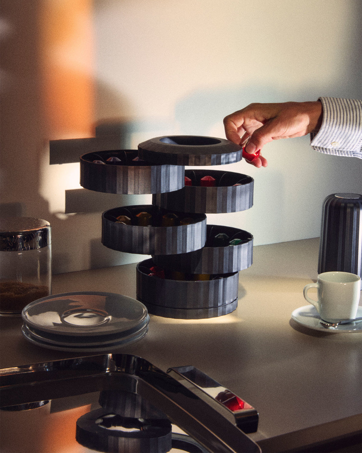Dark grey fluted capsule holder by Alessi, stacked with multiple tiers, as a hand retrieves a red capsule.