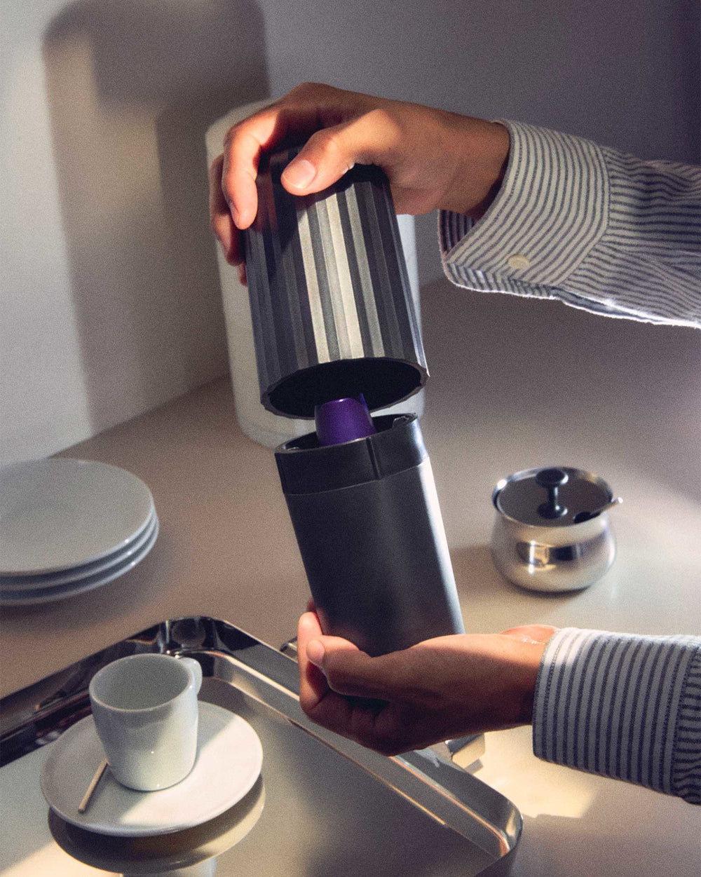 Dark grey capsule crusher by Alessi with a ridged top, held by hands crushing a purple capsule.