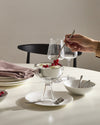 Clear glass and stainless steel dessert bowl by Alessi, filled with ice cream and red currants, with a hand adding more berries.