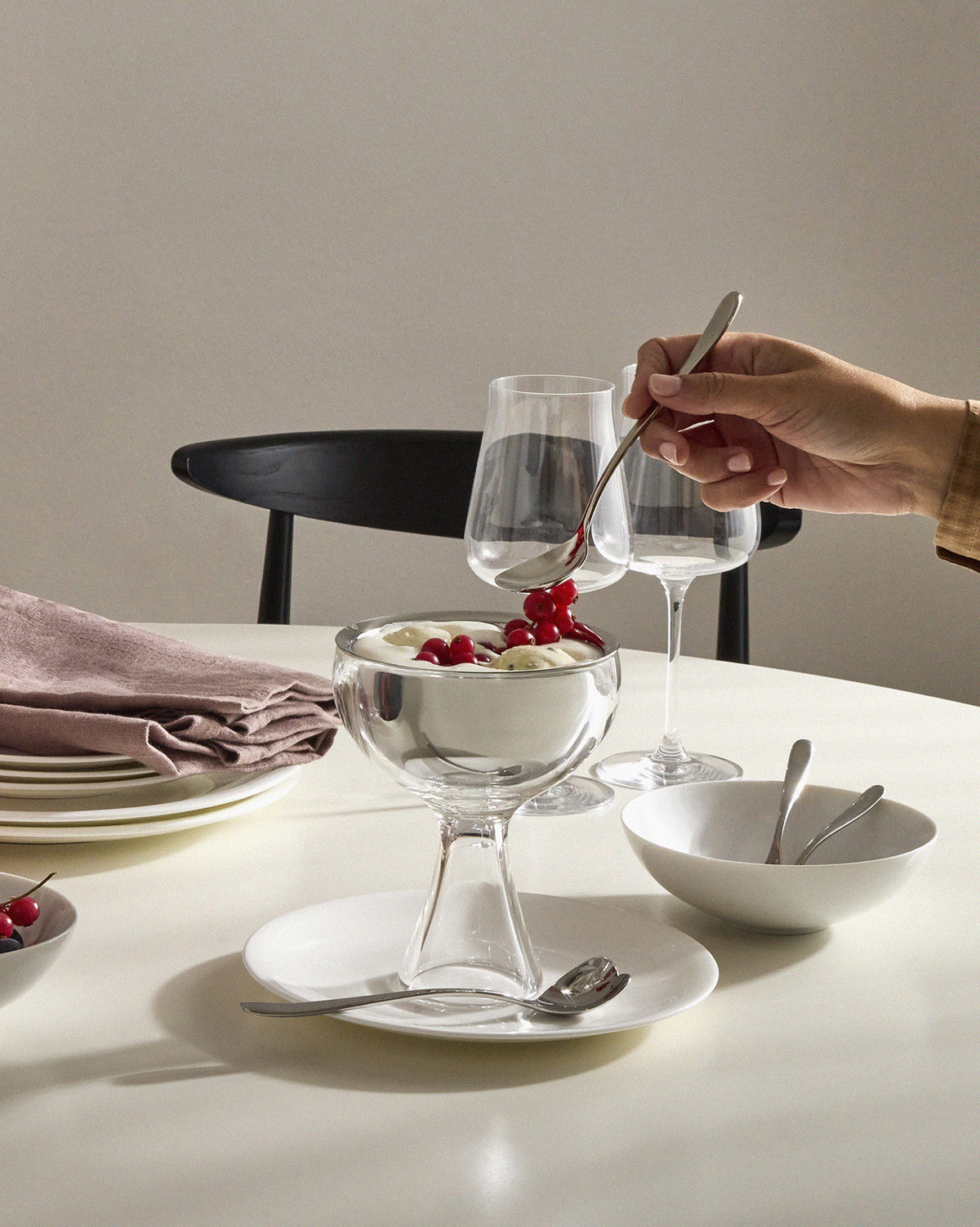 Clear glass and stainless steel dessert bowl by Alessi, filled with ice cream and red currants, with a hand adding more berries.