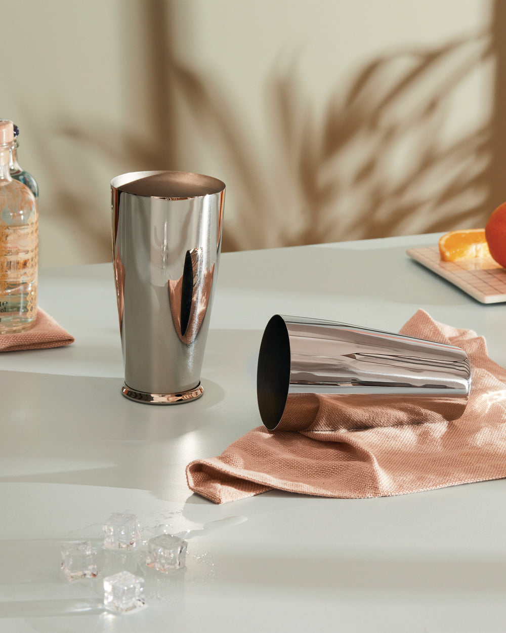 Polished stainless steel and copper cocktail shakers by Alessi, one upright and one lying on a peach textured cloth, with ice cubes.