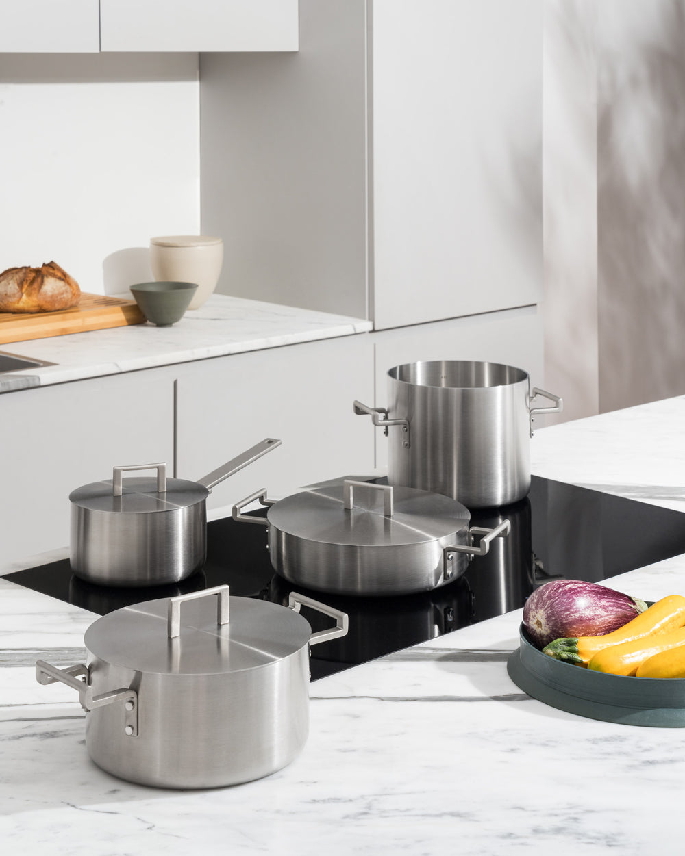Brushed stainless steel lids by Alessi with minimalist square handles, placed on various pots in a modern kitchen.