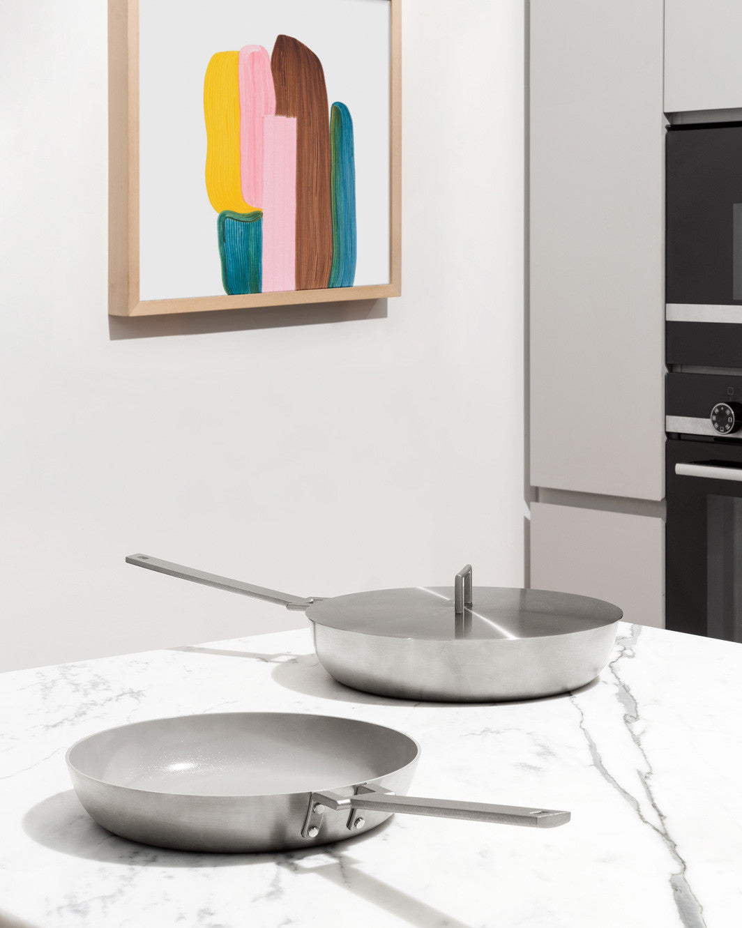 Two silver stainless steel Convivio pans by Alessi, one with a lid, on a white marble kitchen counter.