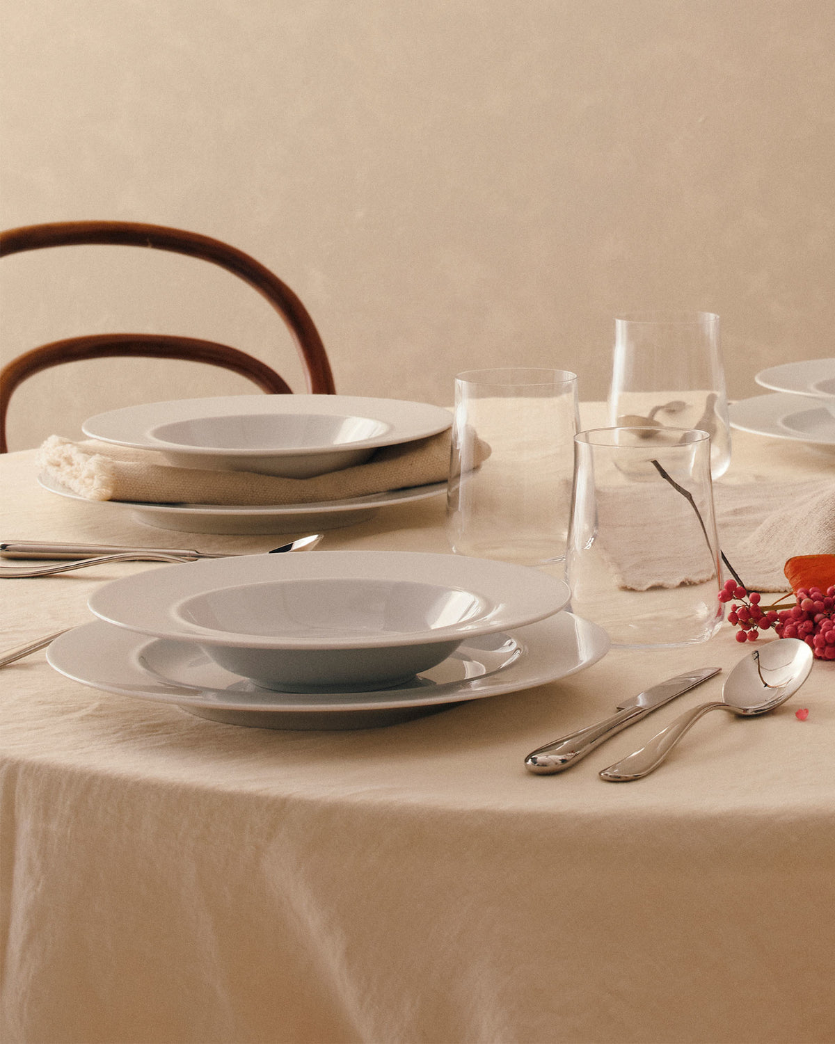 White dining plates by Alessi, stacked with bowls and set on a cream tablecloth with silverware and glasses.