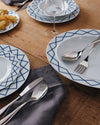 White serving plates by Alessi with blue geometric patterns, on a wooden table with a grey fringed napkin, grapes, cutlery, and a wine glass.