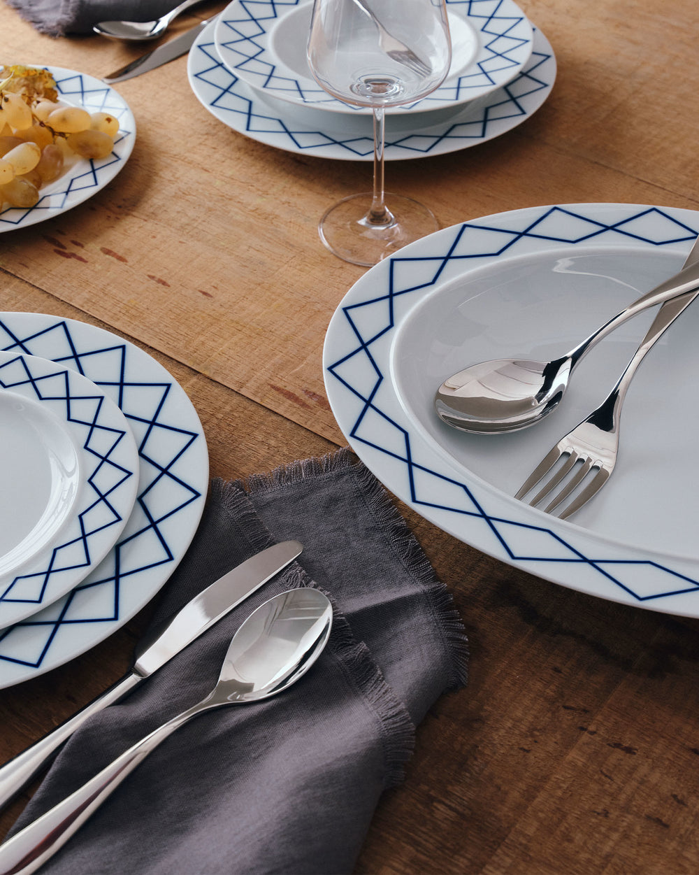 White serving plates by Alessi with blue geometric patterns, on a wooden table with a grey fringed napkin, grapes, cutlery, and a wine glass.
