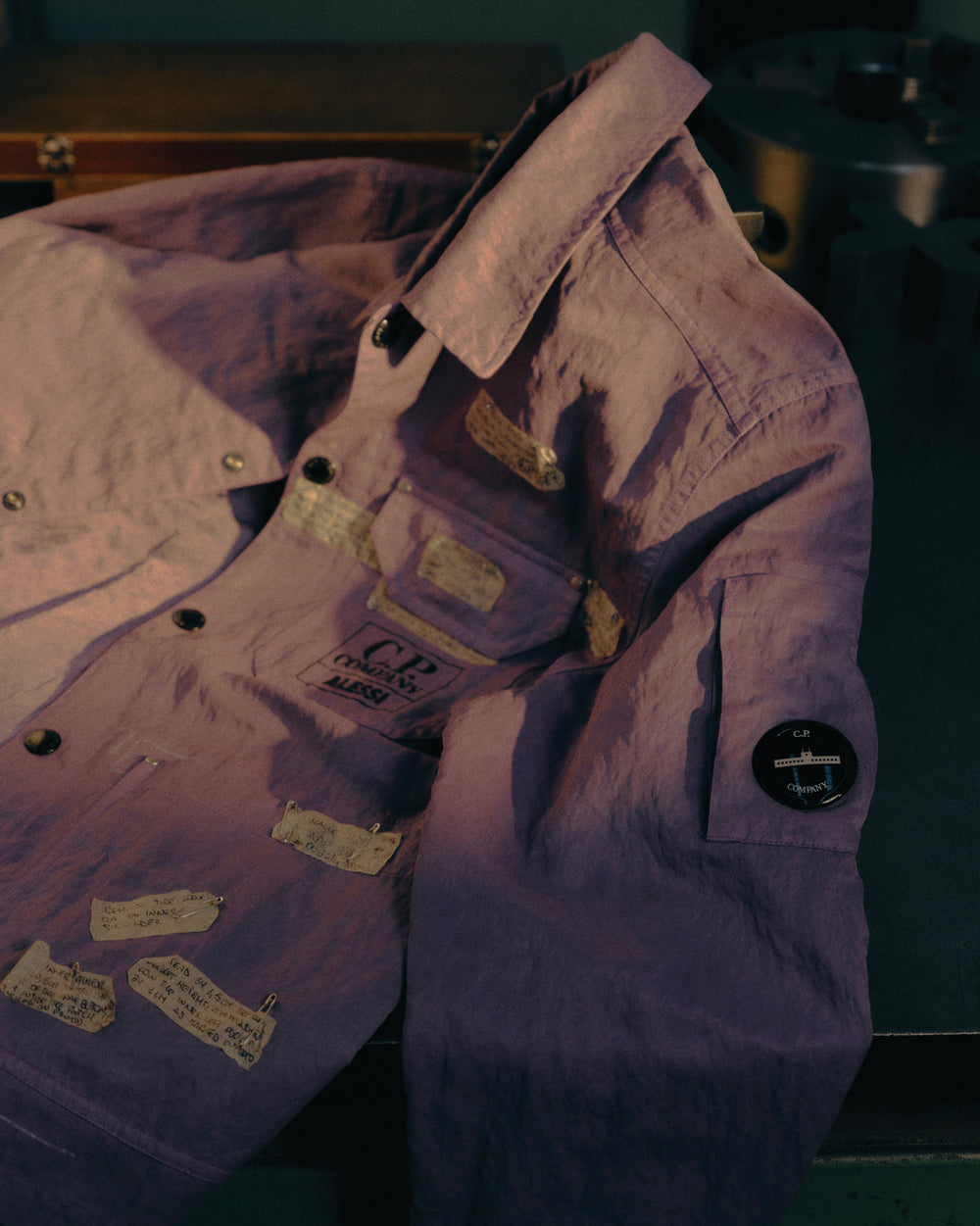 Purple crinkled jacket by Alessi with snap buttons, chest pockets, visible text patches, and a sleeve badge.