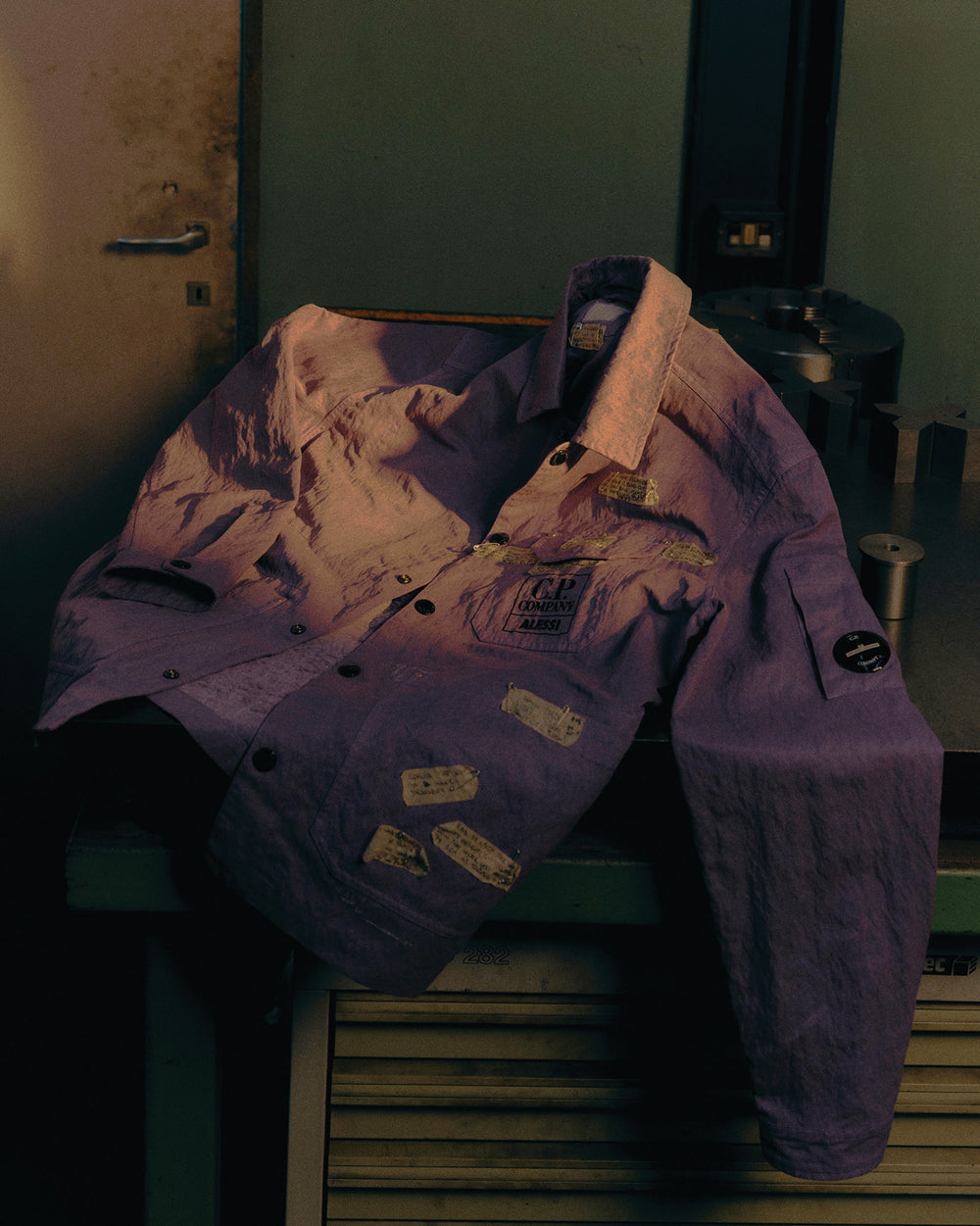 Purple crinkled jacket by Alessi, with 'C.P. COMPANY ALESSI' chest logo, sleeve patch, and decorative text patches.