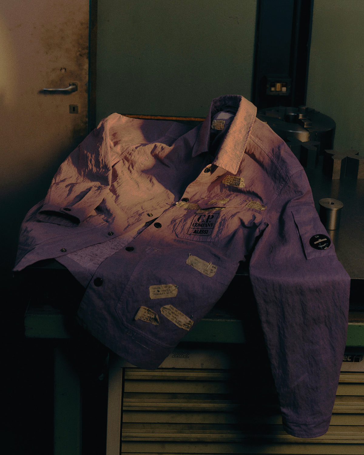 Purple crinkled jacket by Alessi, with 'C.P. COMPANY ALESSI' chest logo, sleeve patch, and decorative text patches.