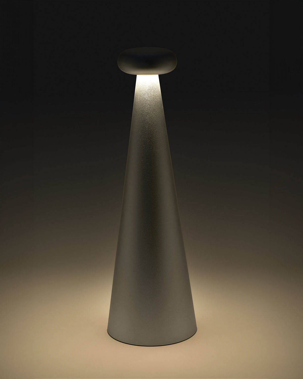 Gray cone-shaped Bedin table lamp by Alessi with a rounded top, emitting warm downward light onto a surface.