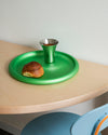 Green round metallic tray by Alessi, holding a matching cup and a pastry on a wooden table.