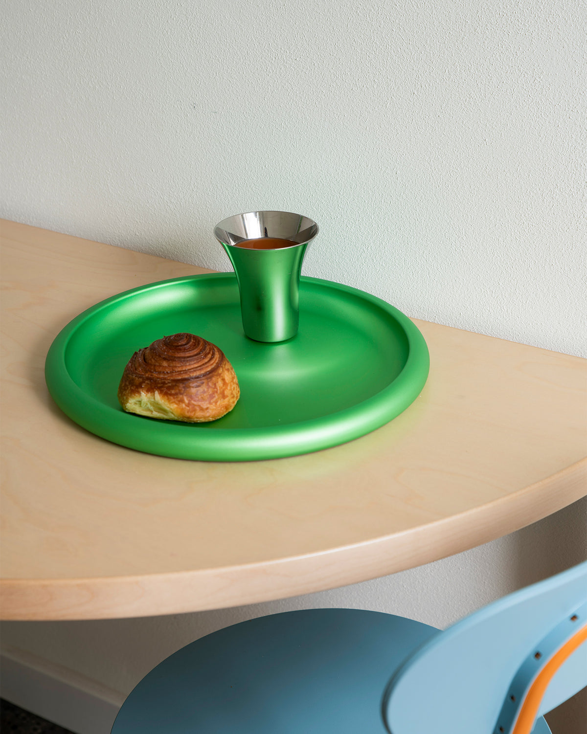 Green round metallic tray by Alessi, holding a matching cup and a pastry on a wooden table.