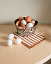 Silver stainless steel bowl by Alessi with a crumpled design, reflecting a brown and white striped napkin, filled with and surrounded by eggs.
