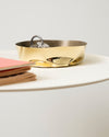 A gold metallic container with a crumpled texture by Alessi, holding silver keys, on a white table next to pink and brown notebooks.