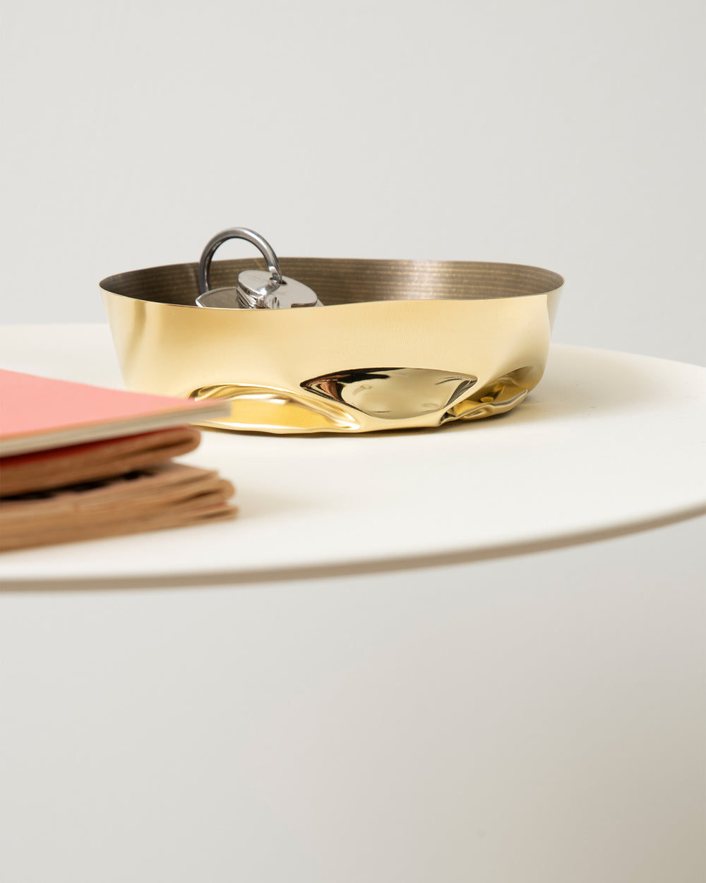 A gold metallic container with a crumpled texture by Alessi, holding silver keys, on a white table next to pink and brown notebooks.