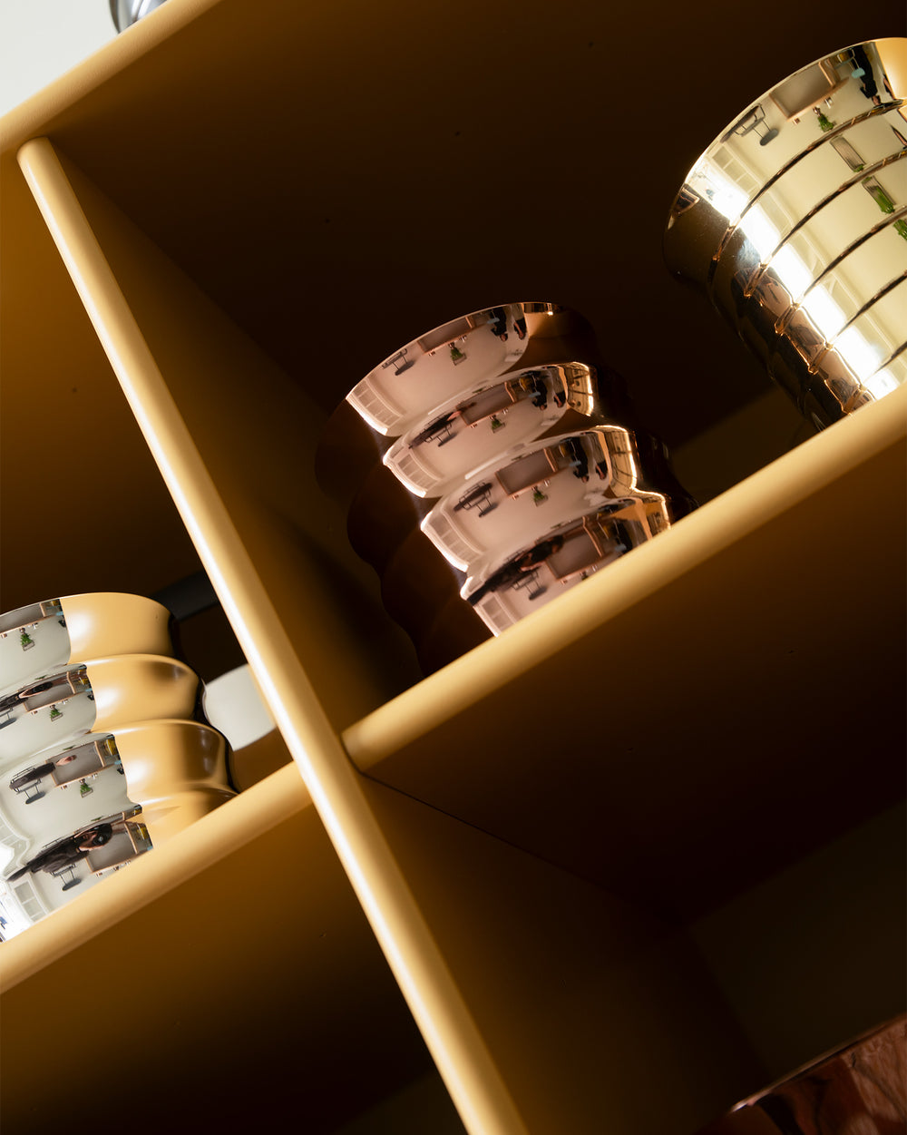 Metallic Dervisci glasses by Alessi, stacked on a mustard shelf, reflecting a room interior.
