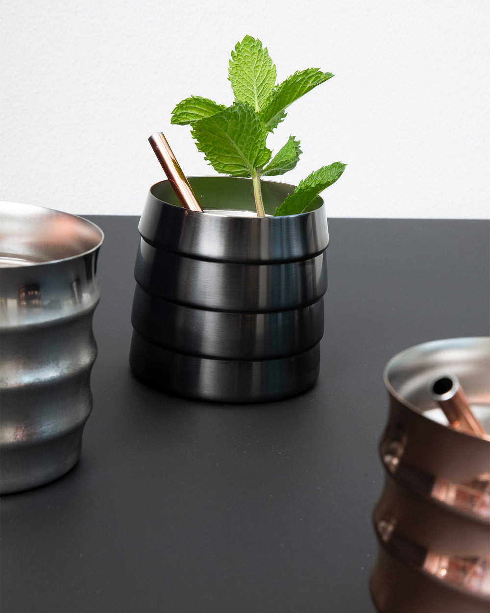 Dark metal tumbler with stacked rings by Alessi, holding mint and a copper straw, beside other metal tumblers.