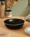 Black pan by Alessi with a silver handle, sitting on a wooden counter with kitchen ingredients.