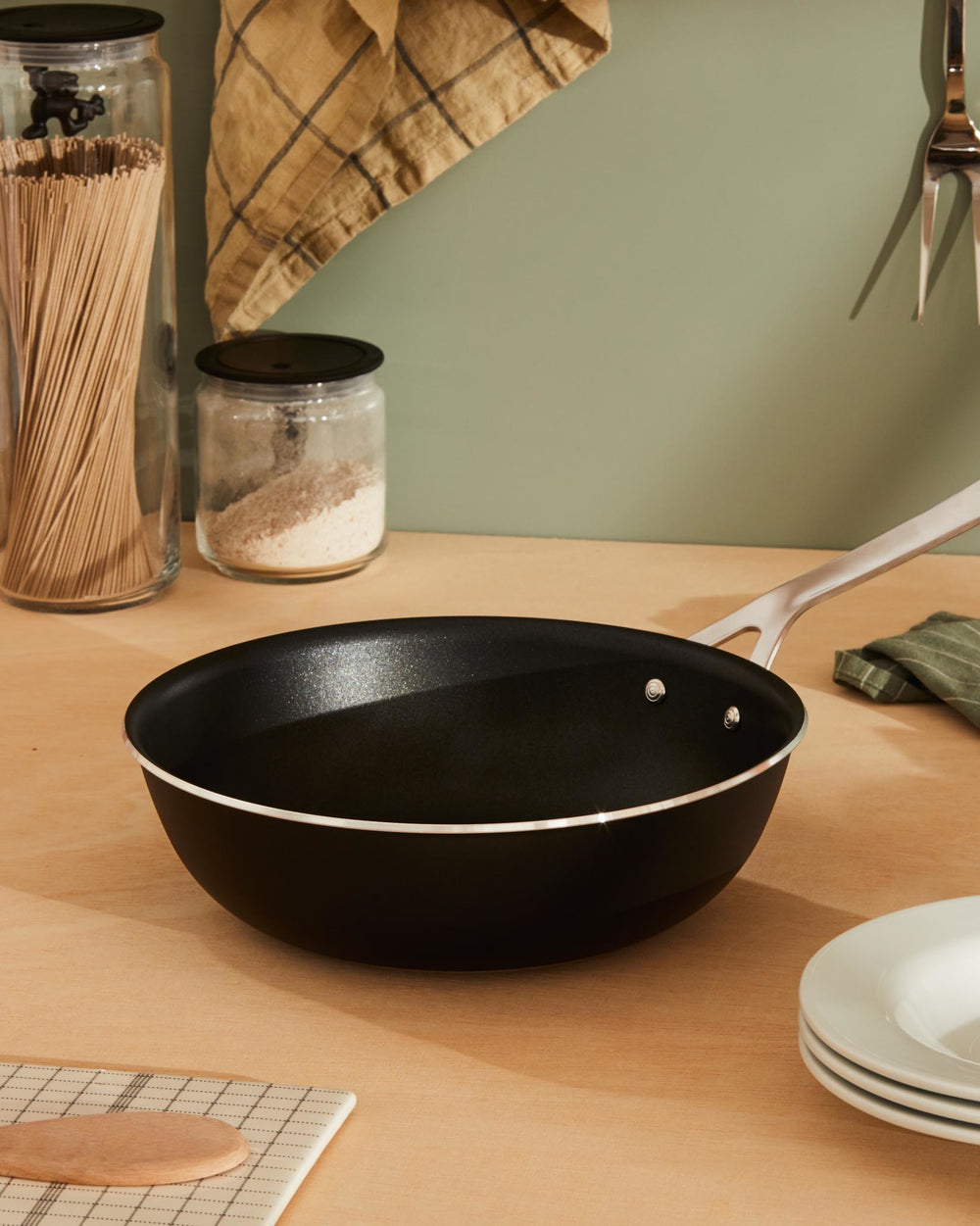 Black pan by Alessi with a silver handle, sitting on a wooden counter with kitchen ingredients.