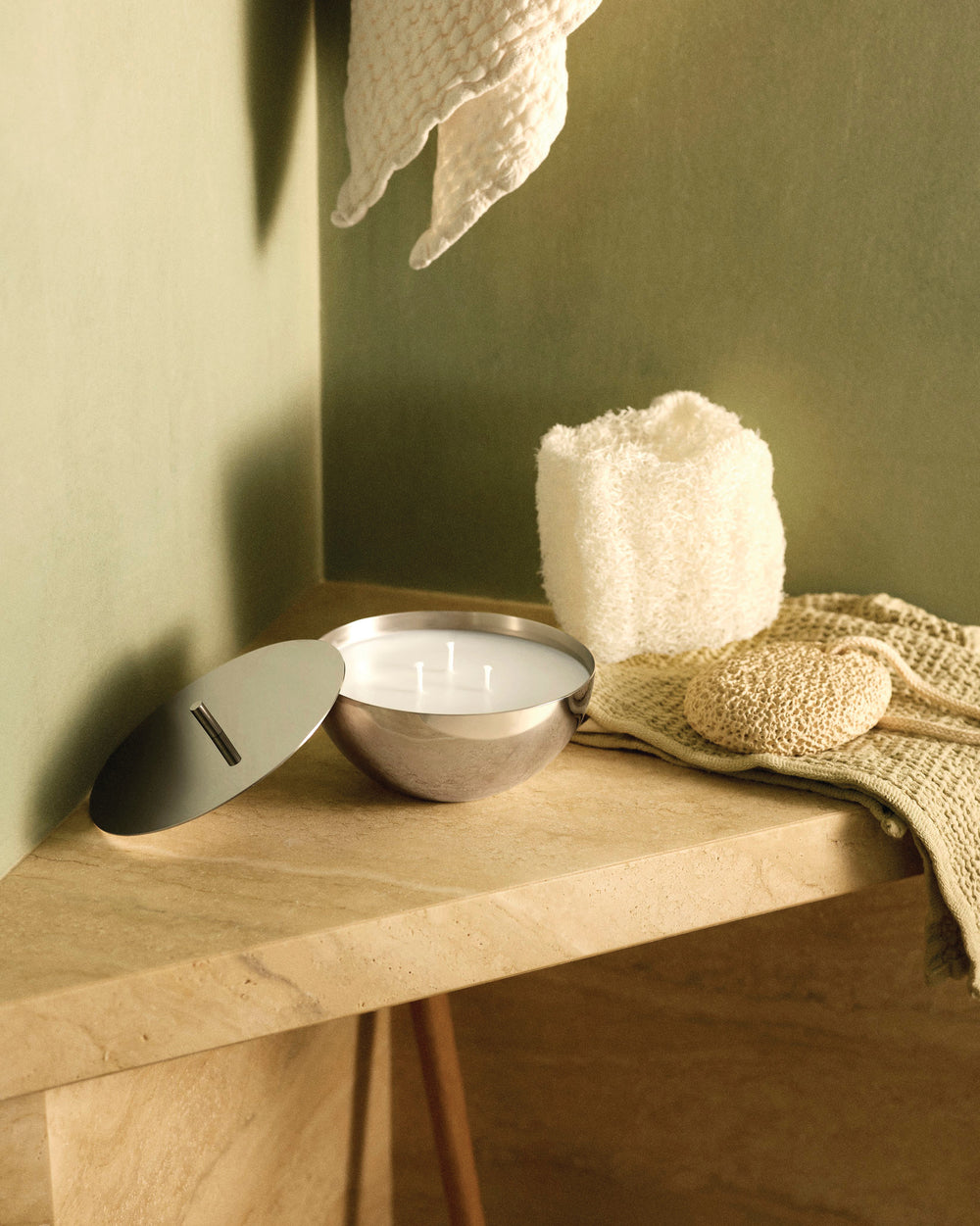White candle in a polished silver bowl by Alessi, with a matching lid, on a beige stone counter.