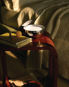 White candle in a polished silver bowl by Alessi, placed on a wooden side table with books and a yellow sphere.