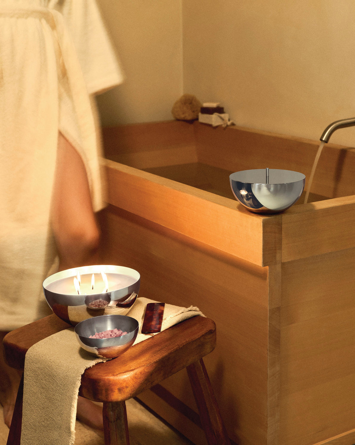 Polished metallic bowl by Alessi, holding lit candles and bath salts, in a warm bathroom setting.