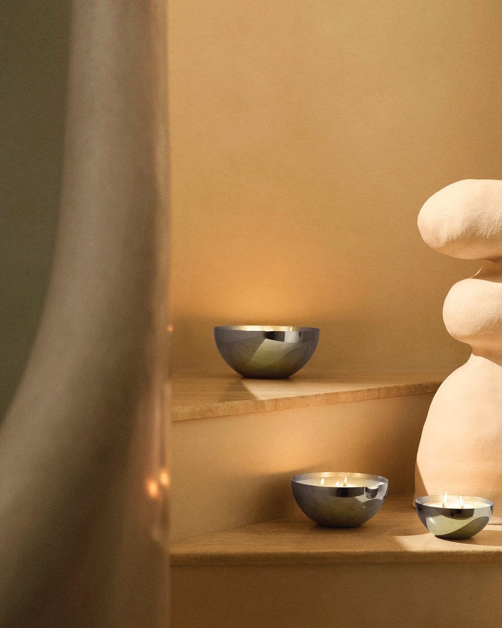 Metallic bowl-shaped candles by Alessi on light wooden steps, reflecting warm ambient light.