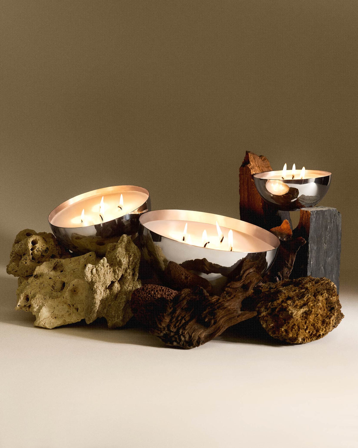 Three silver multi-wick candles in irregular bowls by Alessi, staged with light rocks, dark driftwood, and slate blocks.