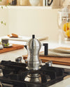 Silver ribbed espresso coffee maker by Alessi, with a black handle, on a gas stove in a bright kitchen.