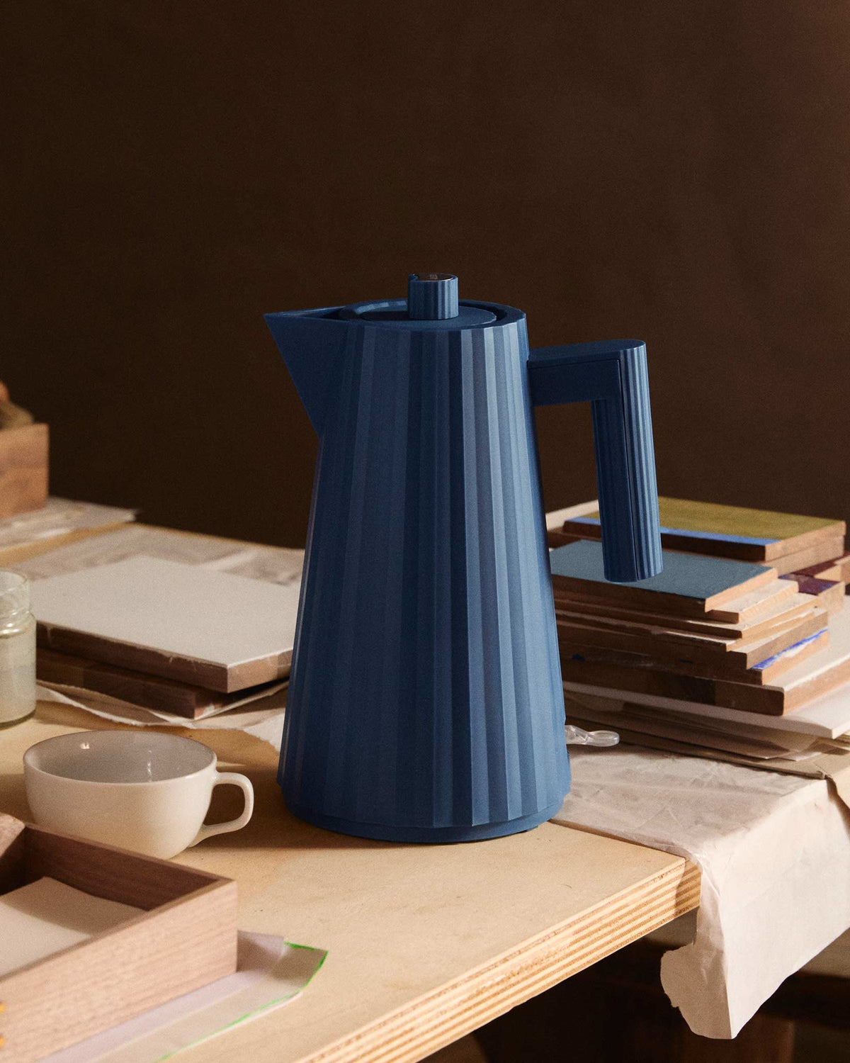 Navy blue pleated electric kettle by Alessi standing on a light wooden table.