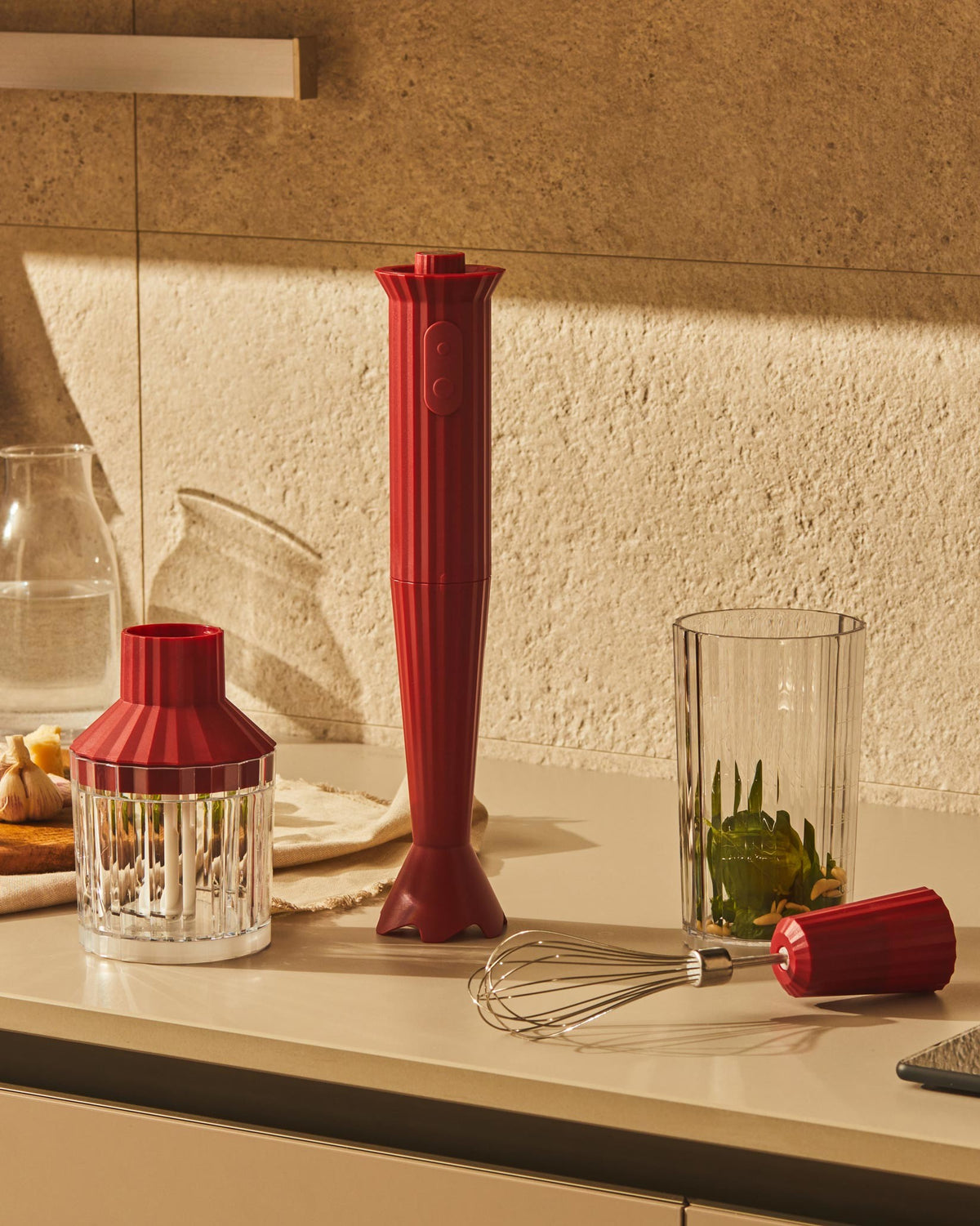 Red ribbed hand blender by Alessi, standing upright with chopper lid, whisk, and pesto ingredients in a glass.
