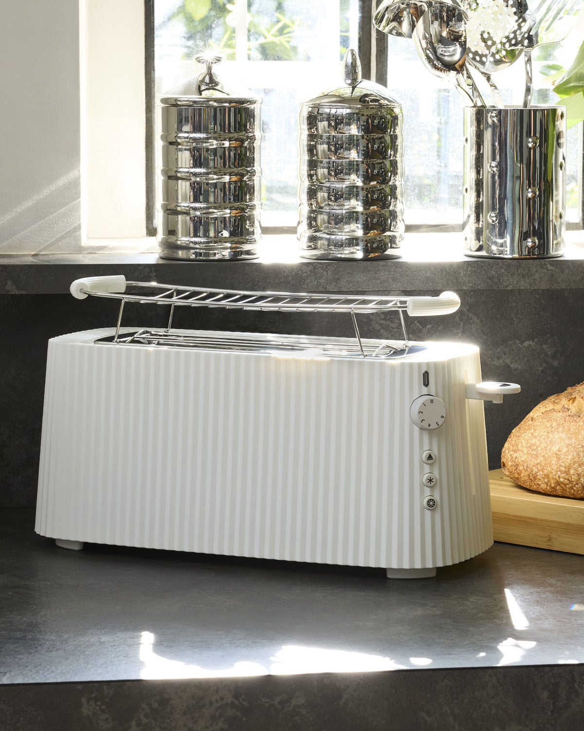 White pleated toaster by Alessi with a chrome warming rack, browning dial, and function buttons on a dark countertop.