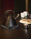 Matte black electric kettle by Alessi with a whimsical bird stopper on its spout and an illuminated digital temperature display.