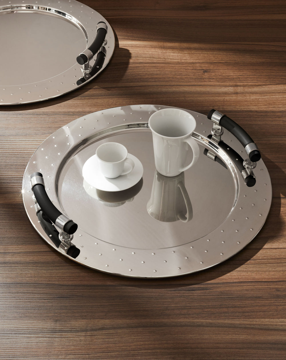 Polished stainless steel serving tray by Alessi with studded rim and black handles, displaying white coffee cups on a wooden table.