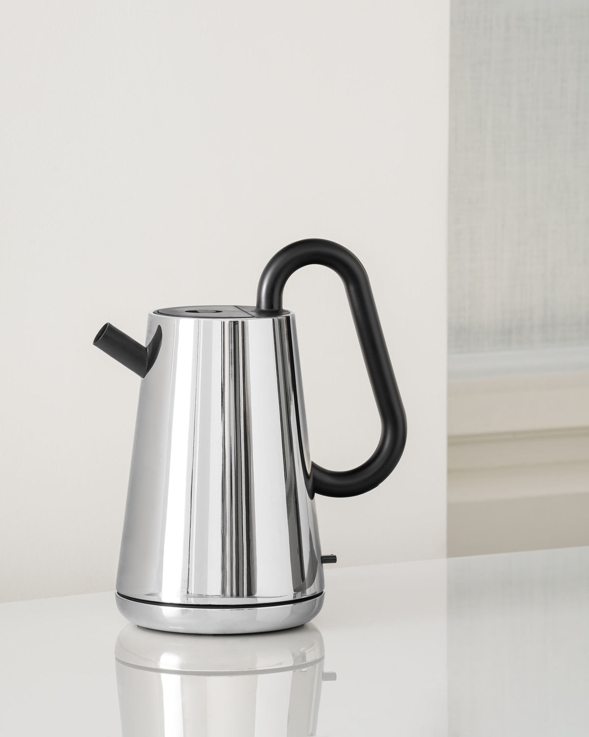 Chrome electric kettle by Alessi with a sleek, modern design, a black looped handle, and a short black spout, on a white reflective surface.