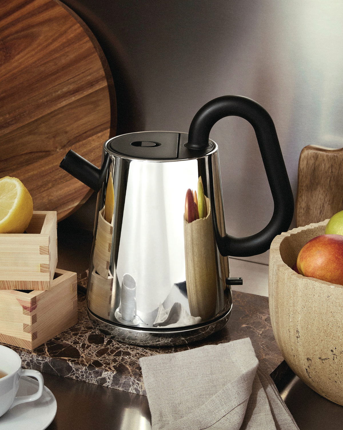 Chrome electric kettle with black handle and spout by Alessi, styled on a marble counter with wooden boxes and fruit.