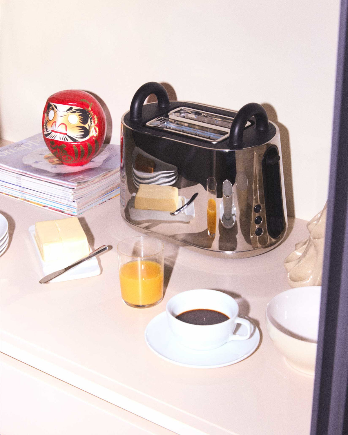 Silver metallic toaster by Alessi with black handles, styled with a red Daruma doll, coffee, and orange juice on a counter.