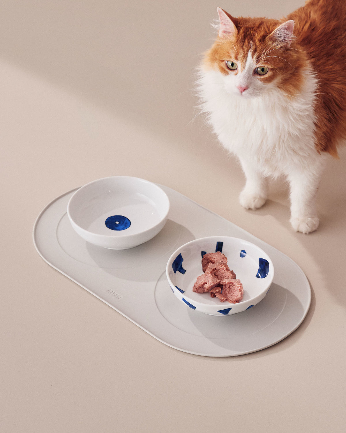 White ceramic cat bowls with distinctive blue patterns by Alessi, presented on a grey feeding mat beside an attentive cat.