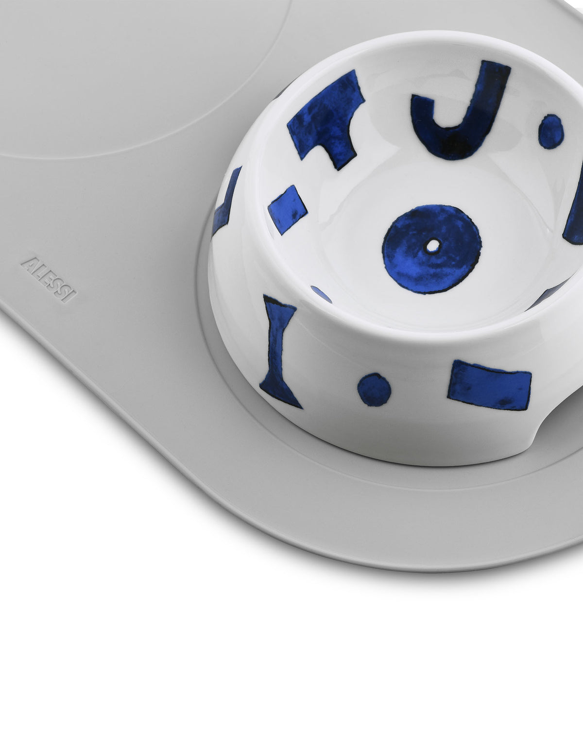 White ceramic pet bowl by Alessi with blue abstract shapes and dots, sitting on a grey silicone mat, close-up.