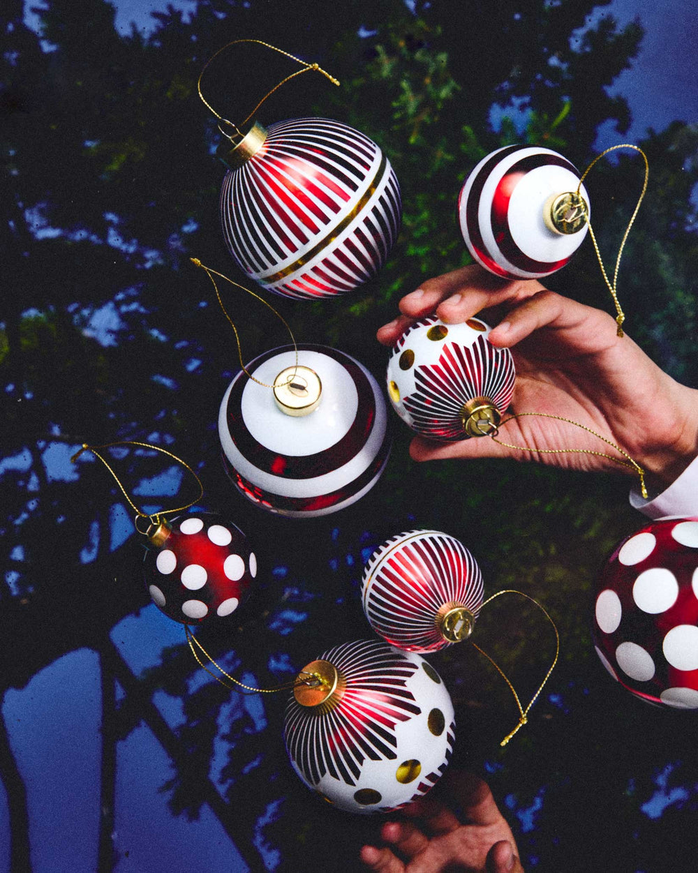 Red, white, and gold Christmas tree ornaments by Alessi with striped and polka dot patterns, one held by a hand.