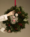 Two diamond-patterned and one star-patterned white and red Christmas ornaments by Alessi, held on a pine wreath with berries.